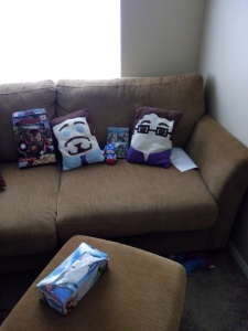 {Image is of a couch. On the couch, from left to right, are an Avengers cereal box, a pillow version of Tony Stark, a tiny stuffed Captain America, the Avengers DVD case, and a pillow version of Bruce Banner. In the foreground, on an ottoman, is a box of tissues.}
