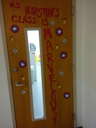 {Image is of a classroom door decorated with replicas of Captain America's shield, Thor's hammer, and Iron Man's mask. Each replica has a child's first name on it. Red lettering on the door reads, "Ms. Kirstin's Class Is Marvelous".}