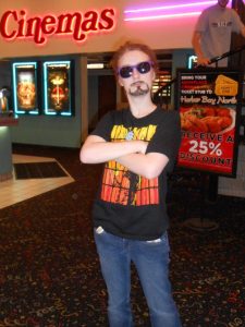 {Image is of me cosplaying as Tony Stark for the Iron Man 3 premiere. I am wearing a black Iron Man t-shirt from the boys' section of Target and purple sunglasses I got for free somewhere. I have an arc reactor button pinned to my shirt. My hair is dyed brown, and I've painted a goatee on my face with mascara. All of this outfit was decided on just hours before the movie started.}