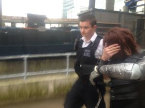 {Image is of a police officer leading the cosplayer away. The cosplayer is handcuffed. He is shielding his face with his non-metal hand.}